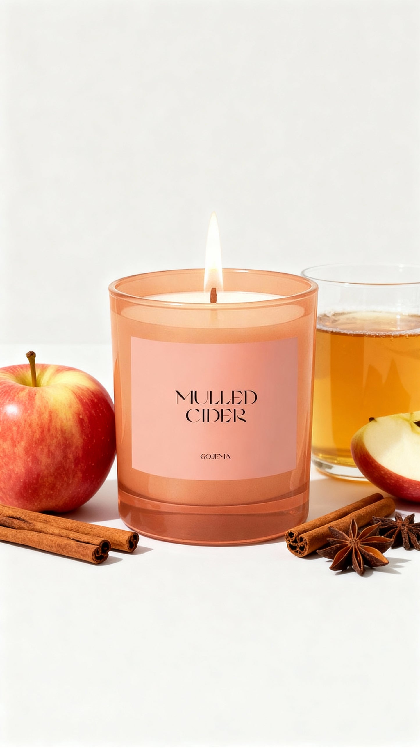 Pink 'Mulled Cider' candle with an apple, cinnamon stick, and glass of mulled cider on a white background
